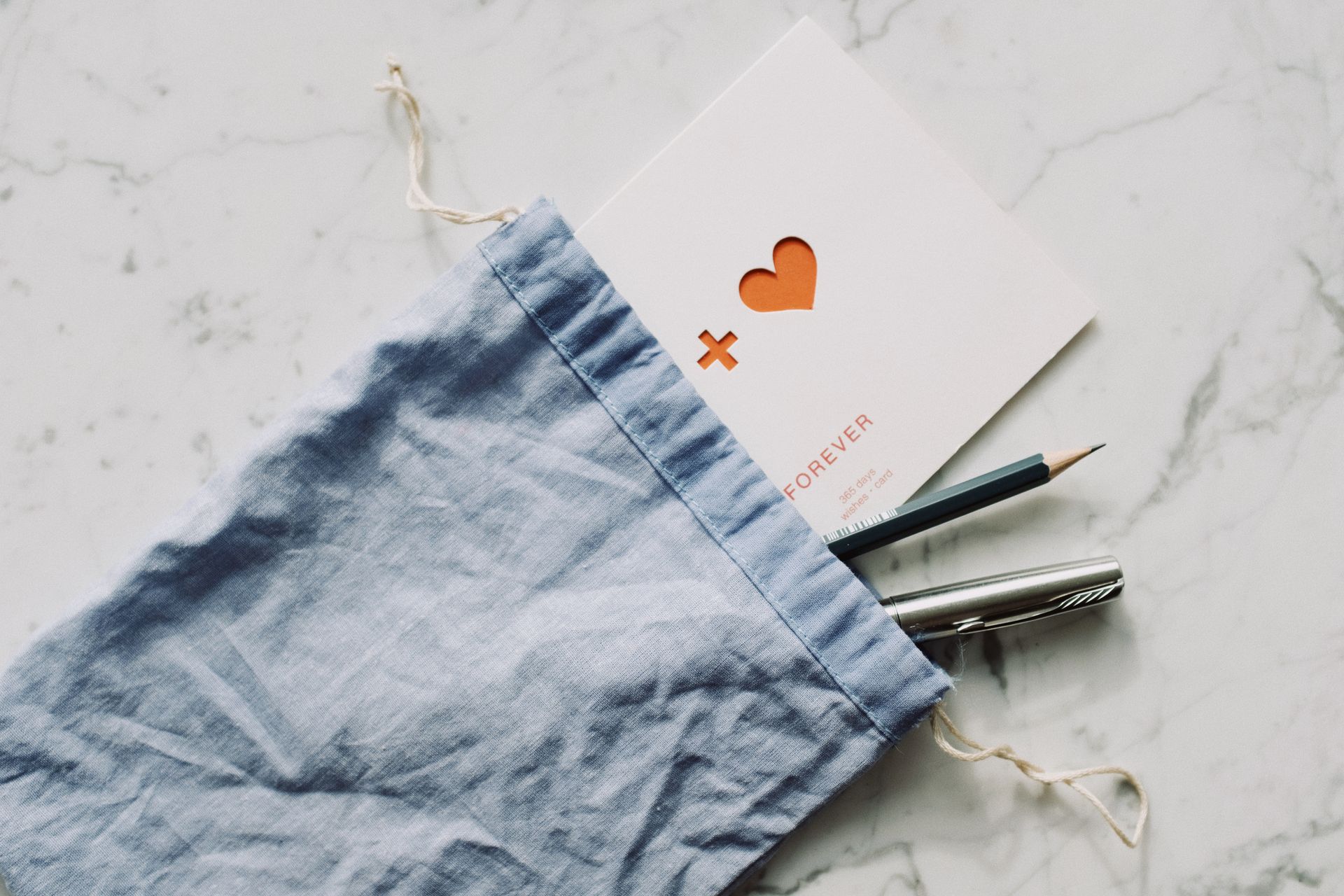 From above view of white postcard with red drawn heart with pen and pencil in blue textile bag placed on marble table in room