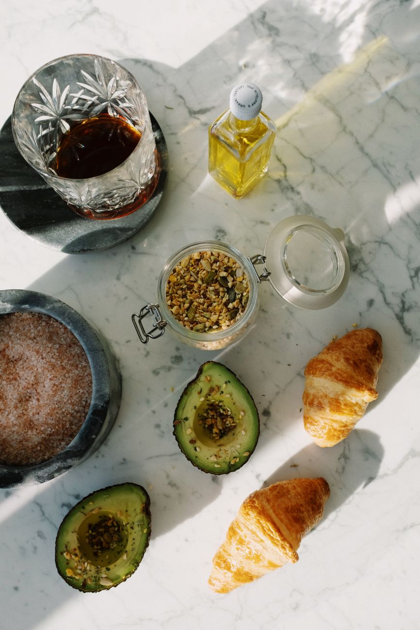 Top view of delicious croissants and halves of fresh tasty avocado sprinkled with various seeds placed near pink salt with seed mixture in glass jar and other dressing ingredients arranged on white marble counter