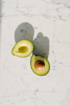 High-angle view of two avocado halves on a marble surface, featuring natural shadows.