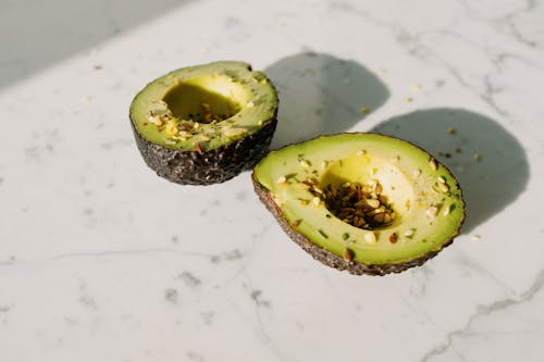 From above of green halved avocado sprinkled with various seeds placed on white marble counter served for healthy vegetarian breakfast preparation