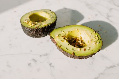 Free From above of green halved avocado without kernel sprinkled with various seeds placed on white marble desk served for healthy vegan breakfast preparation Stock Photo