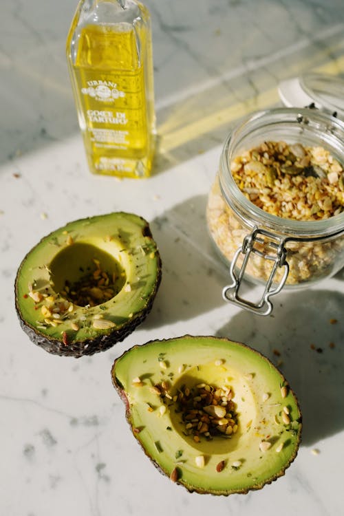 From above composition of fresh avocado halves sprinkled with seed grains from glass jar of seed mixture placed near bottle of natural oil on white marble table
