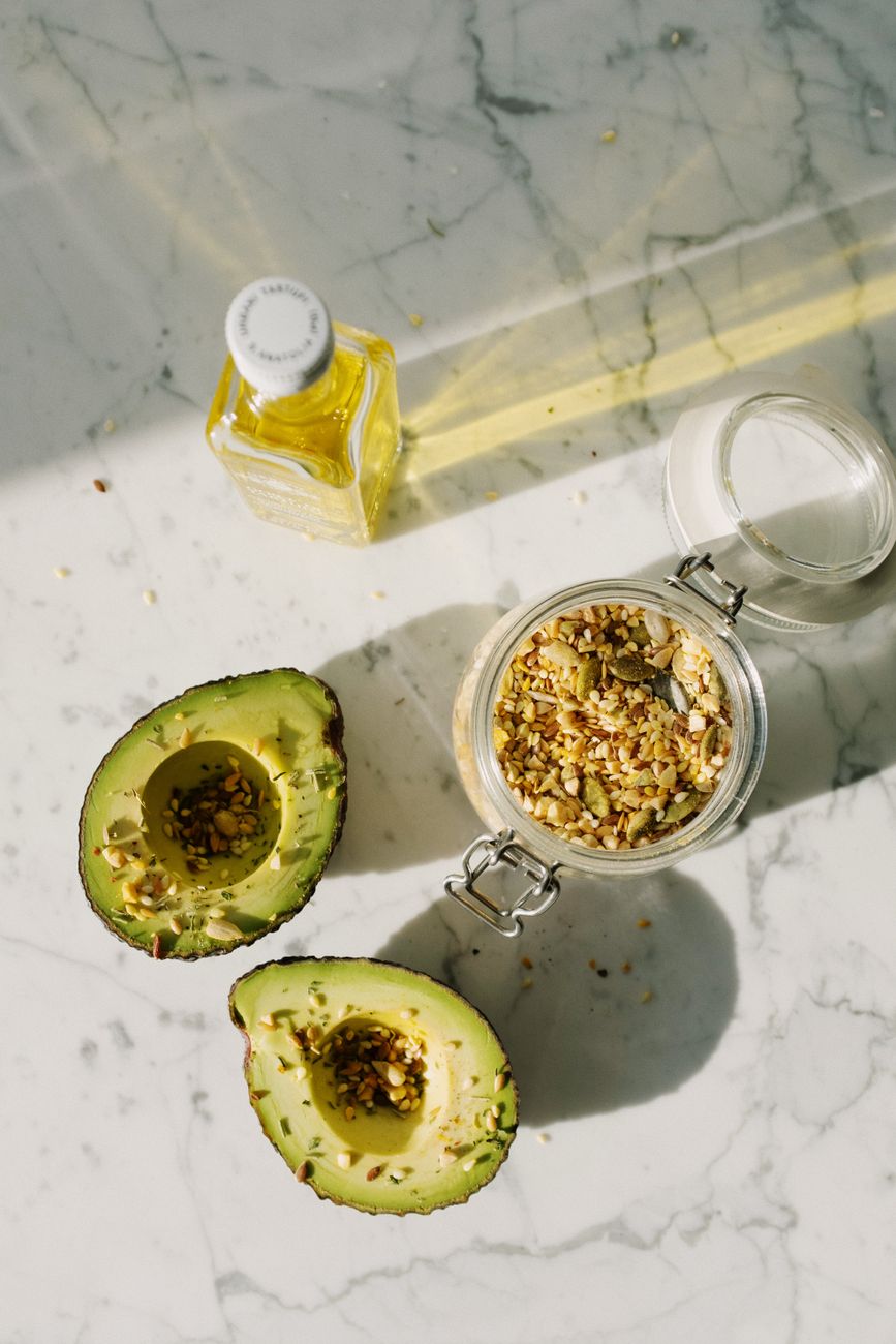 Top view composition of halved avocado without kernel sprinkled with various seeds and glass container of seed grain mixture placed near oil glass battle on white marble table