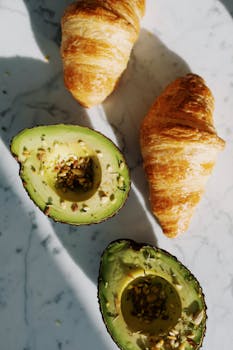 Delicious fresh croissants and ripe avocado halves on a marble countertop with natural lighting.