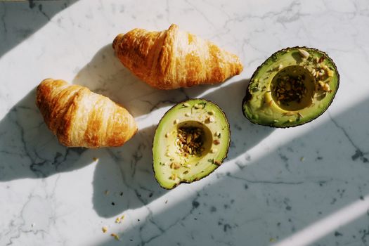 A delicious morning breakfast featuring croissants and avocado halves with seeds on a marble surface.