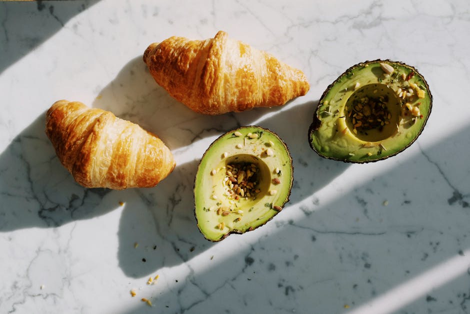 Avocado Green vs Birch Natural: Organic Cert Showdown A delicious morning breakfast featuring croissants and avocado halves with seeds on a marble surface.