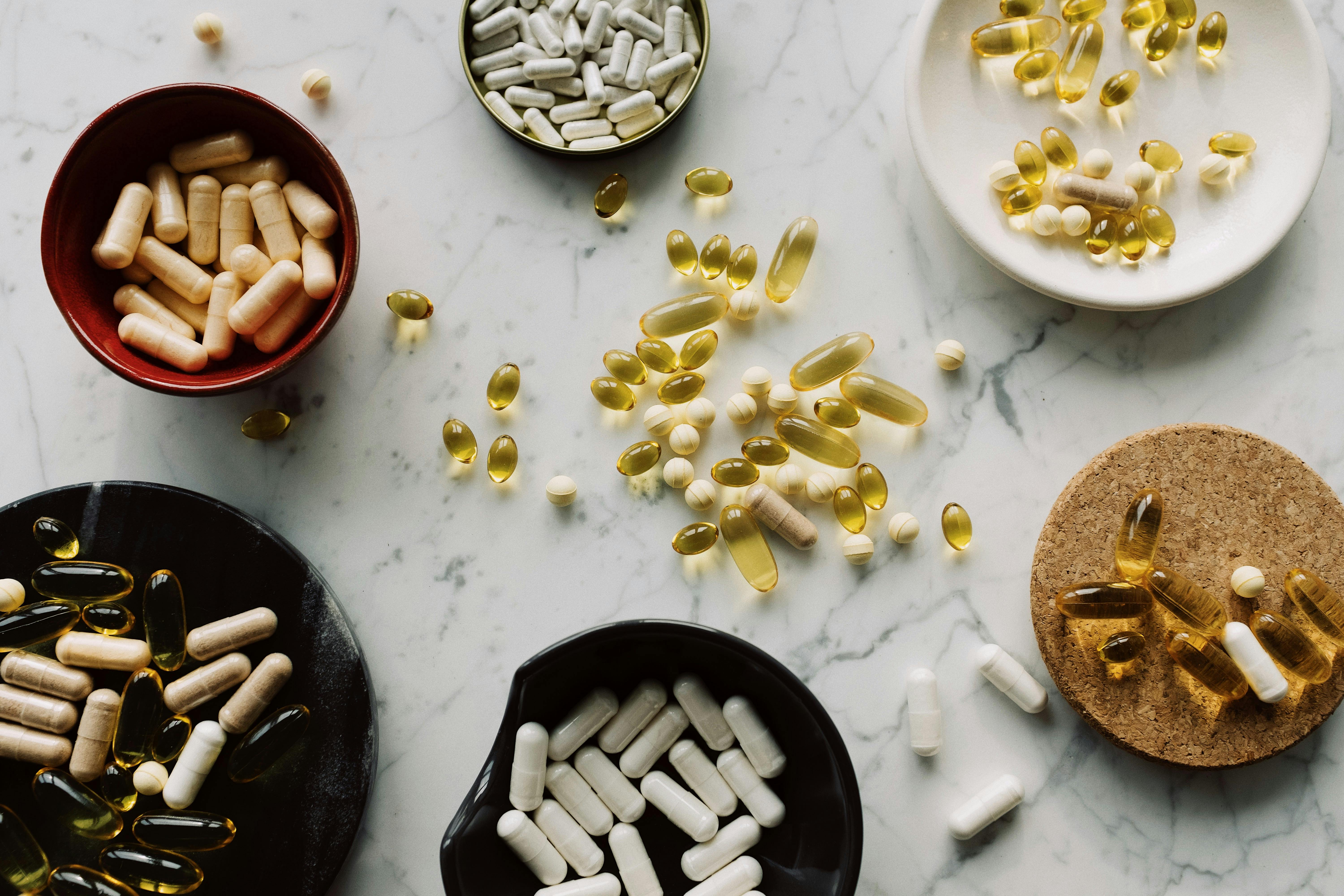 Various pills and capsules displayed in bowls and scattered across surface, including tablets of different sizes and colors.