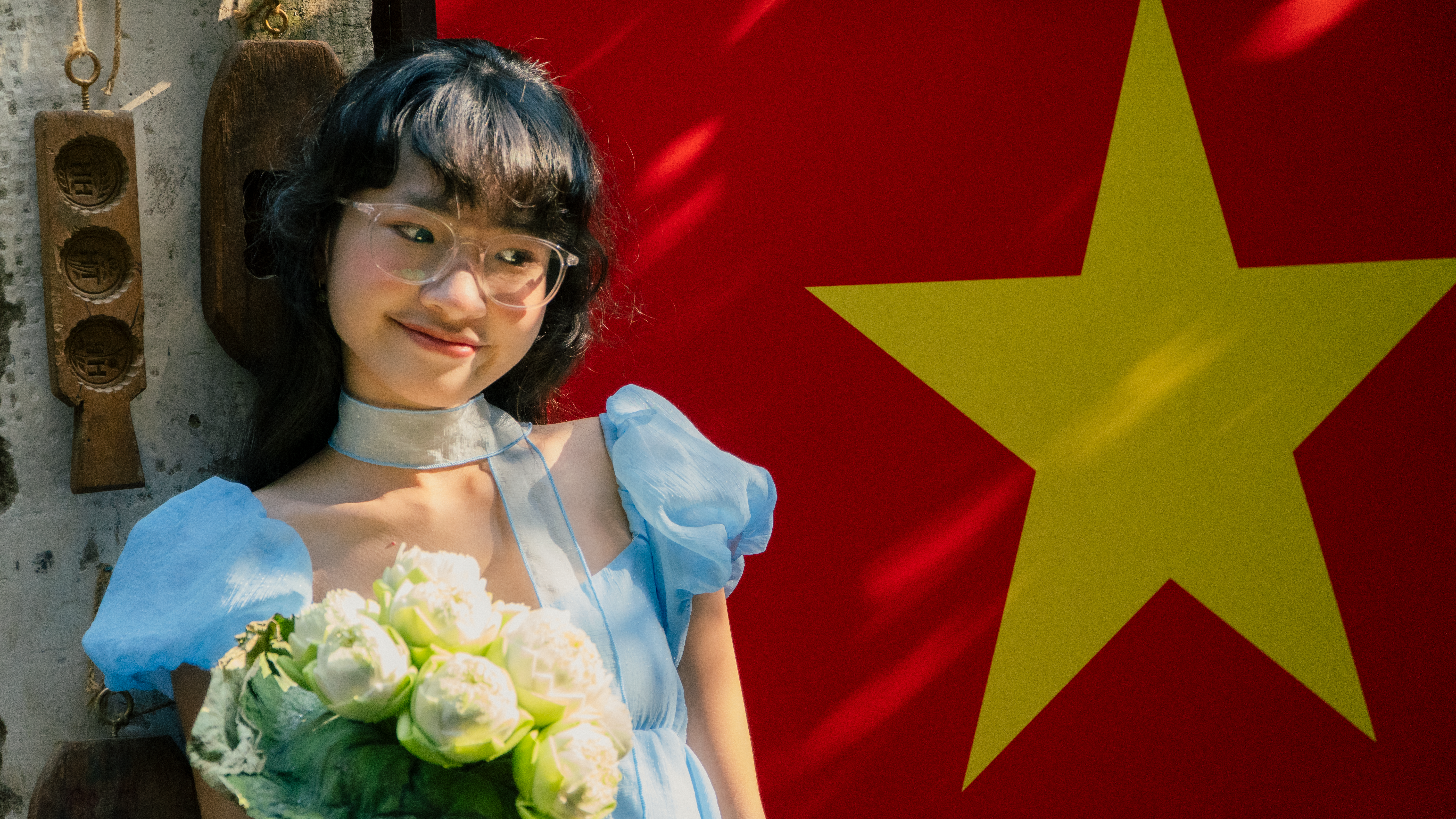 Young woman wearing blue dress and glasses smiling beside a Vietnamese flag.