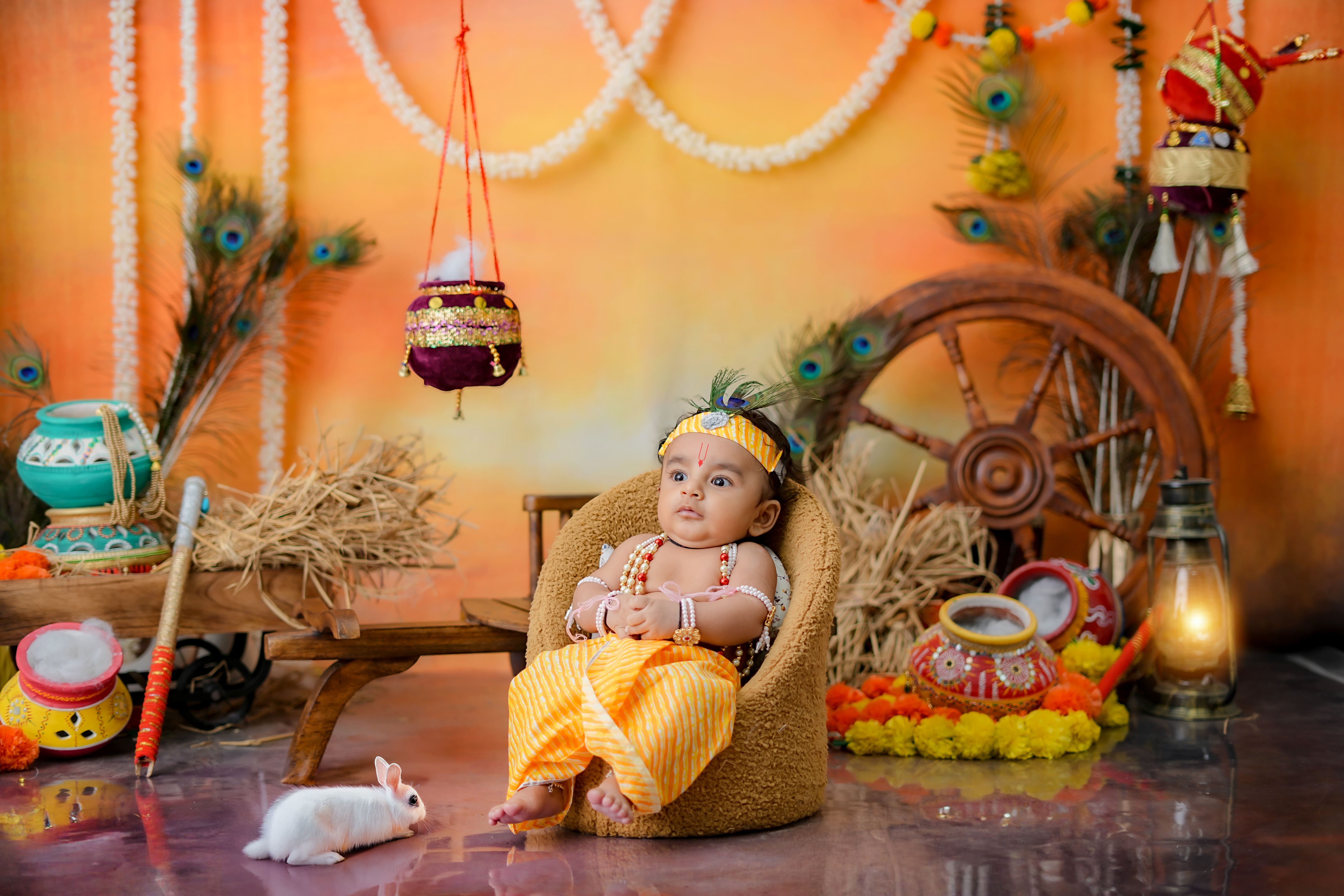 A cute baby sits in a vibrant Indian festival backdrop with decorations and a rabbit.