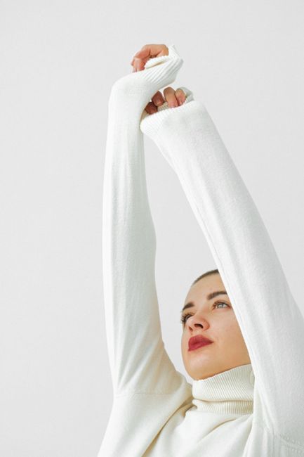 Woman in white turtleneck raises arms, set against a minimalist white background, exuding calm and reflection.