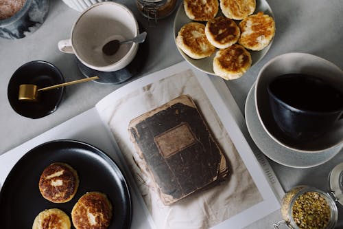 Free Top view of plate with delicious curd pancakes placed on recipe book surrounded by various plates on gray tabletop in kitchen Stock Photo