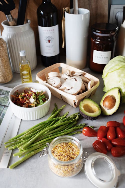 Free From above of box with chopped mushrooms avocado cut in half tomatoes fresh asparagus placed on table near plate with greenery mix and bottle of oil for making salad on table Stock Photo