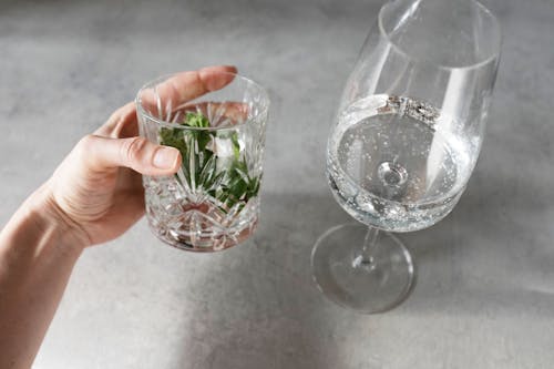 Crop person holding glass with herbs and water near wineglass with tonic on gray table