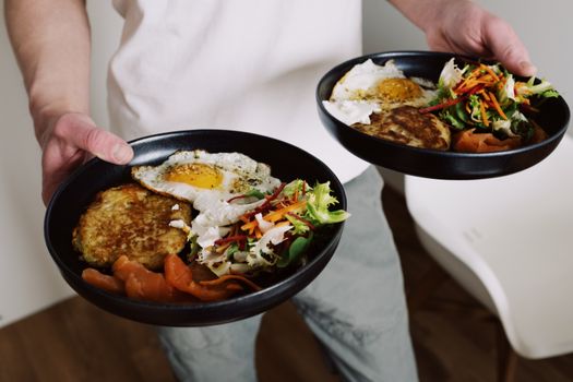 Faceless male in white clothes holding black plates with fried eggs and fresh salad for breakfast in cozy kitchen