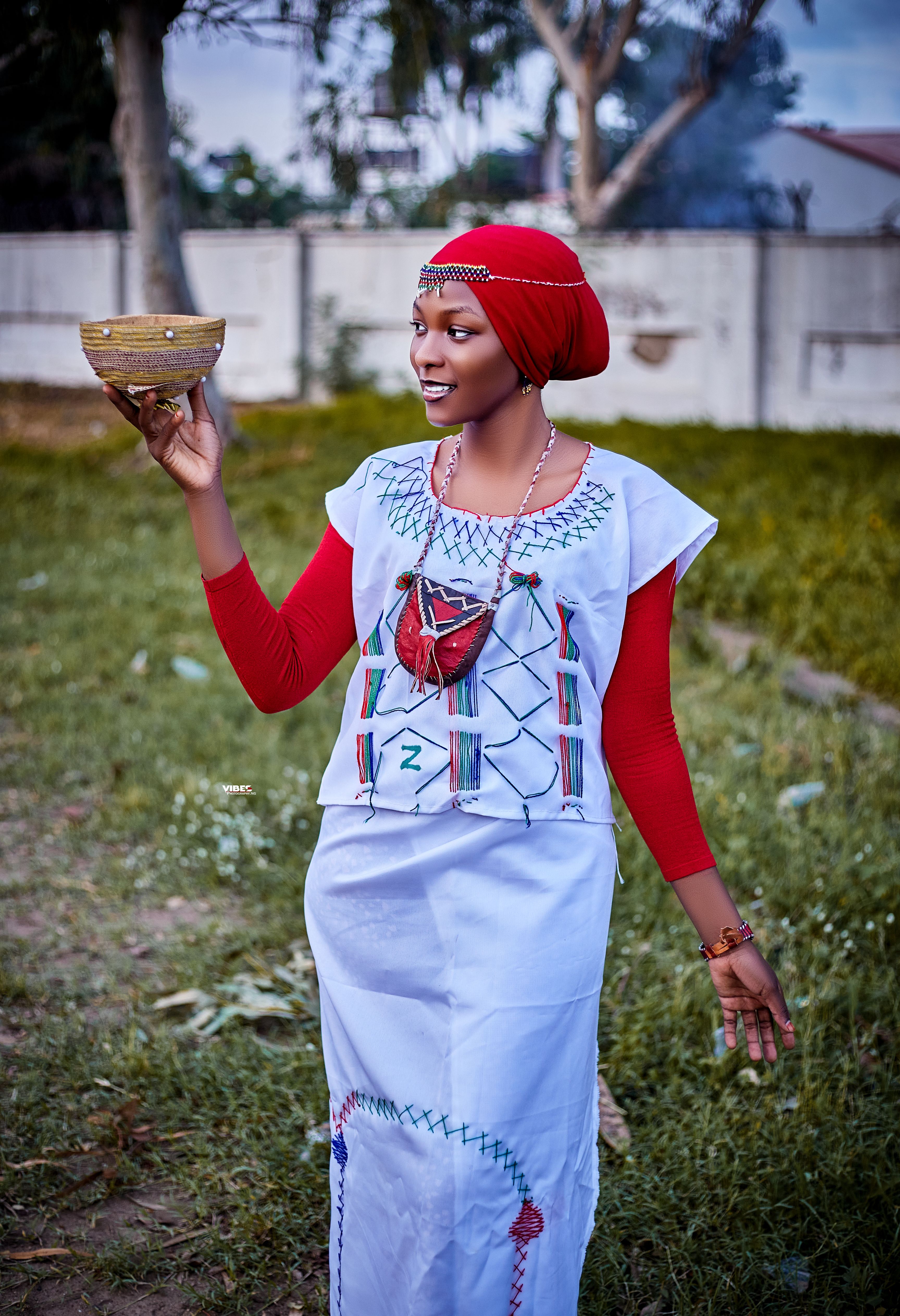 Portrait of a smiling woman in traditional African attire holding a woven bowl outdoors.