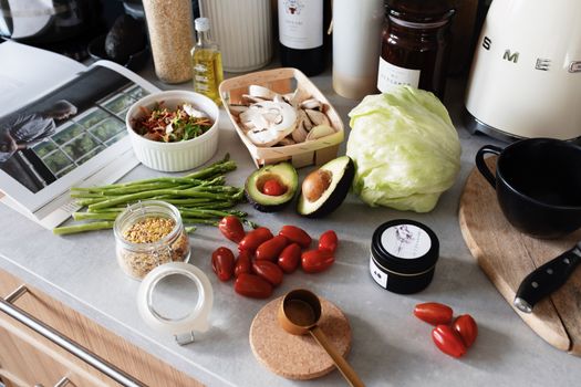 From above of various organic vegetables chopped ingredients near cutting board kitchenware appliances and trendy magazine placed on gray countertop in kitchen at home