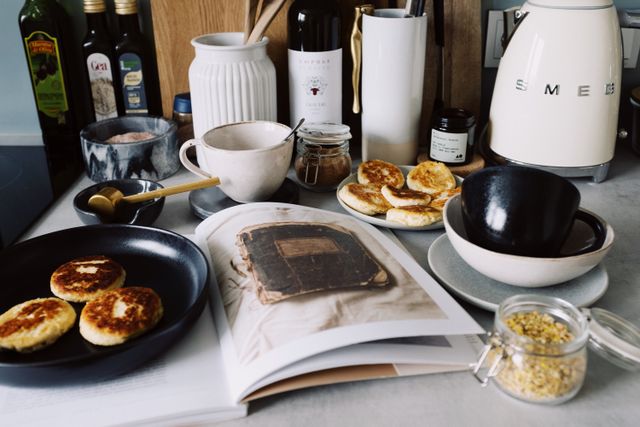 One portion of homemade cheese pancakes in frying pan and another on plate locating near opened stylish magazine among kitchen utensils and appliances in bright kitchen at home