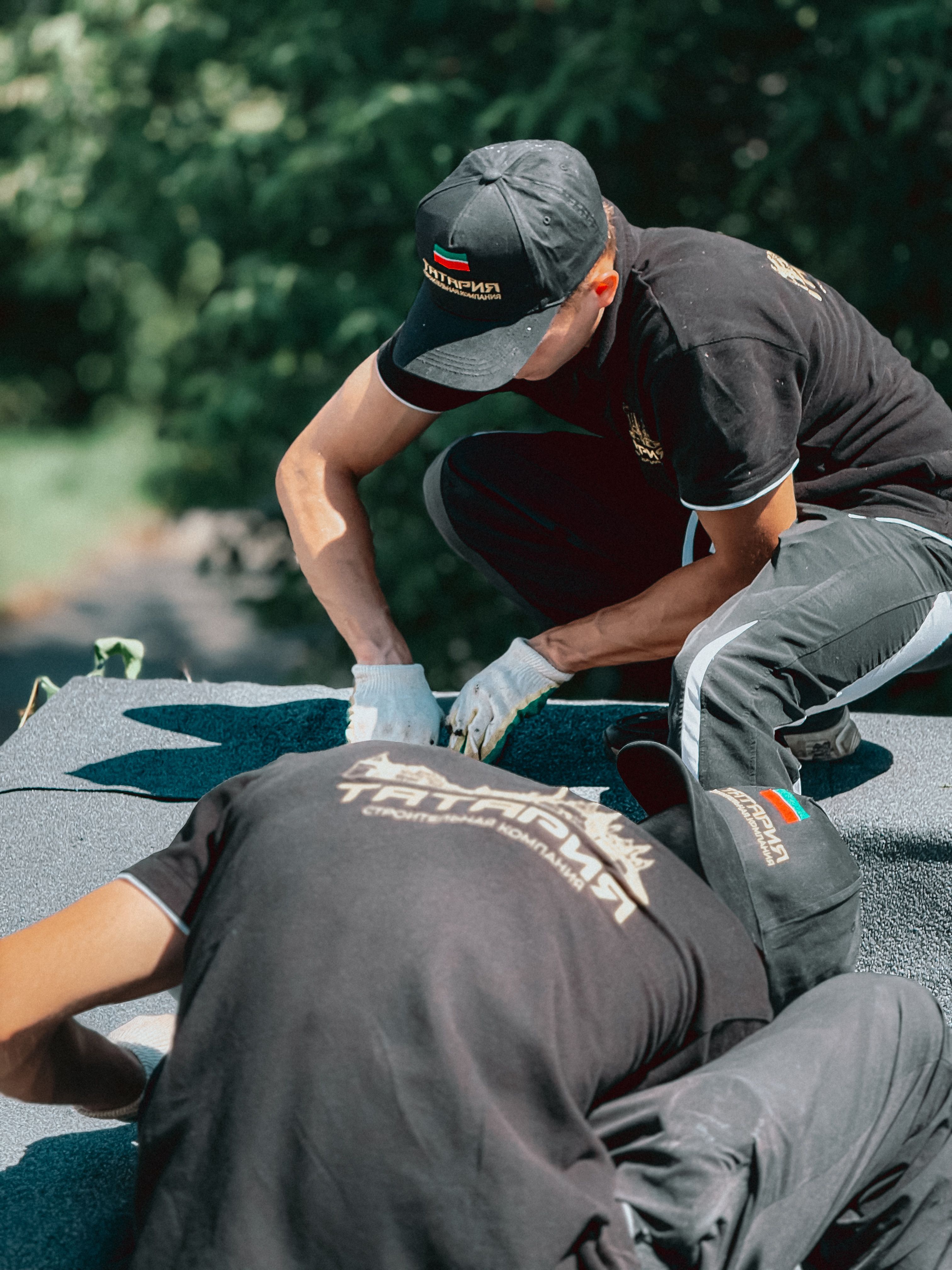 Two workers applying roofing material on a building roof. Outdoors, sunny day.