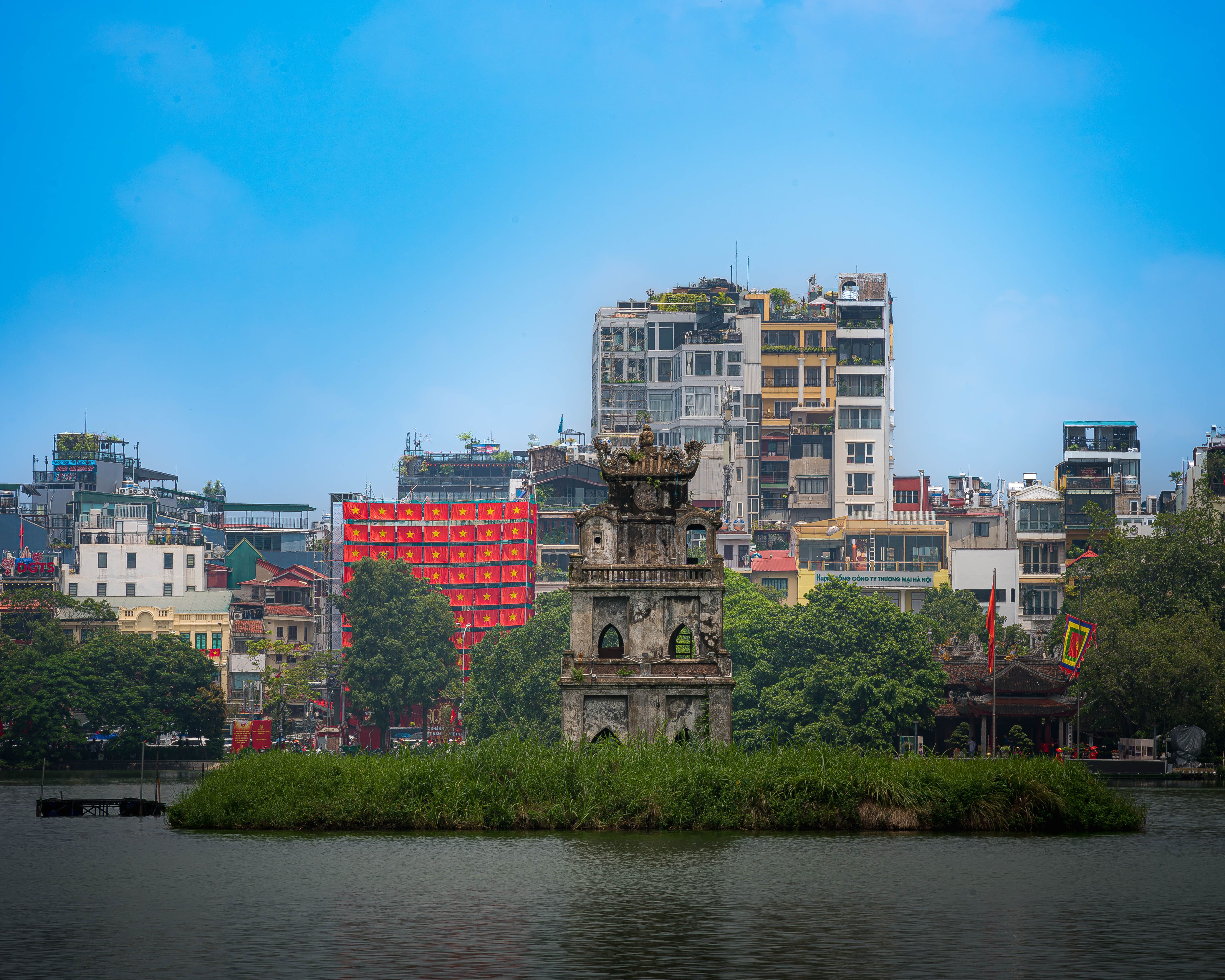 Turtle Tower standing prominently in Hoan Kiem Lake, Hanoi, with cityscape background under a bright sky.