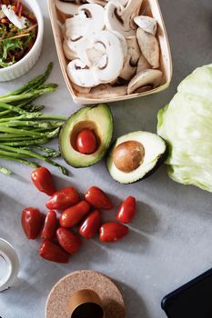 Top view of juicy red cherry tomatoes two halves avocado green asparagus chopped champignons in wicker basket head of cabbage near kitchen utensils and bowl with salad on gray table