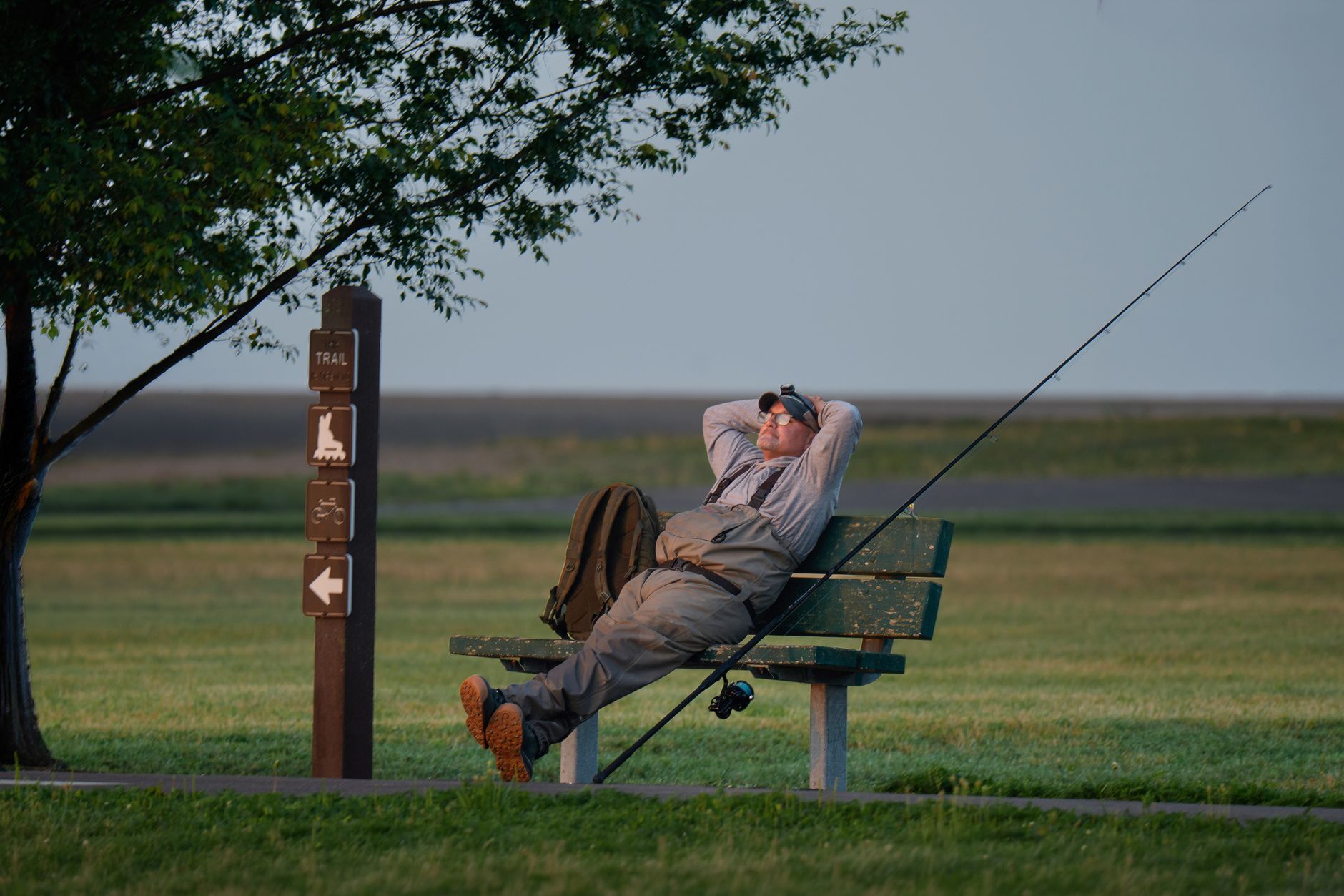 A man rests on a park bench under a tree with fishing equipment, enjoying a tranquil outdoor moment.