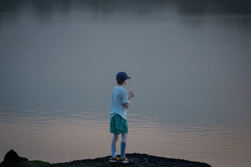 Free Young boy stands on lakeshore at sunrise, creating a tranquil and reflective moment. Stock Photo
