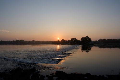 Free Serene sunrise at a waterfront with gentle waves and distant trees, creating a tranquil ambiance. Stock Photo