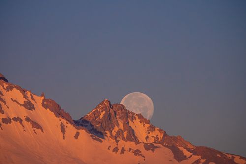 Free Full moon rising over a snow-capped mountain peak during sunset, creating a dramatic natural scene. Stock Photo