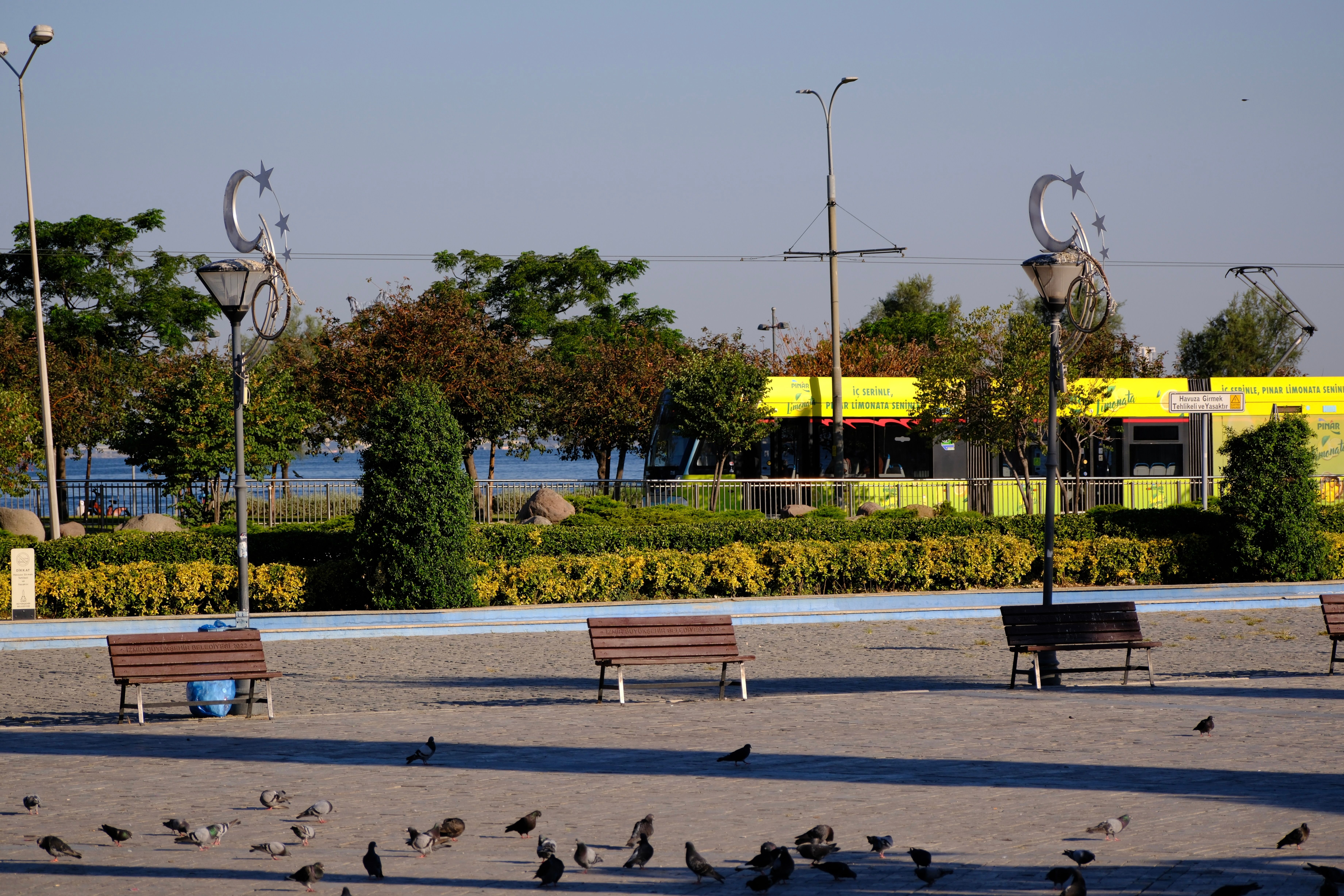 Peaceful park view in İzmir with benches and tram passing by, captured in early morning light.