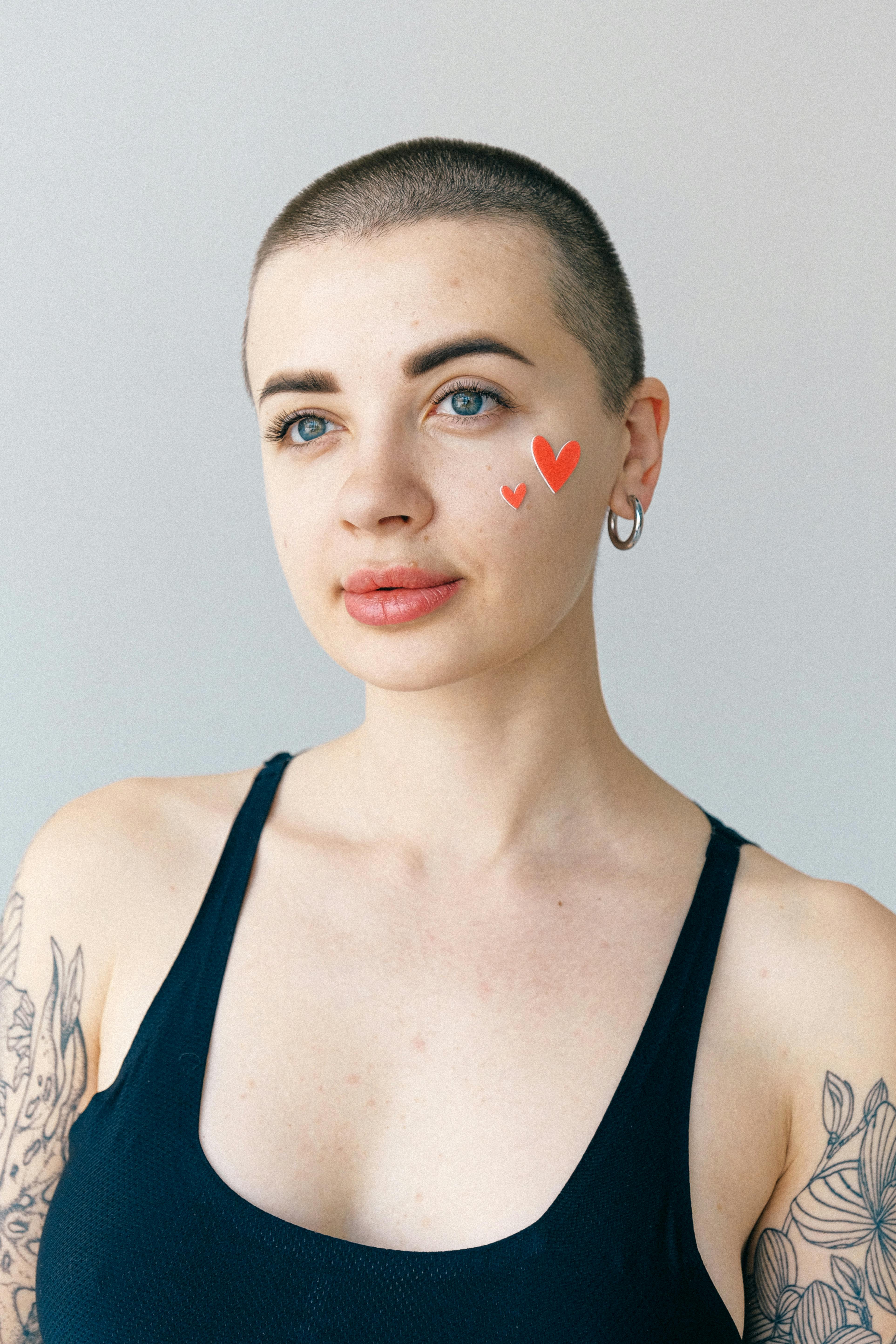 Stylish tattooed woman with shaved head and heart design makeup on her cheek in a minimalist setting.