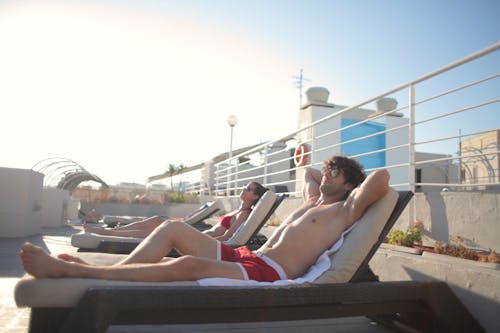 Side view of young man and woman in swimsuits and sunglasses relaxing on sun loungers together and enjoying summer sunny day during vacation