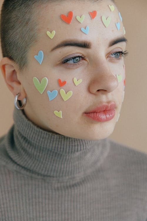 Crop pensive lady in turtleneck with short haircut and multicolored stickers in form of hearts on face standing on beige background in studio and looking away