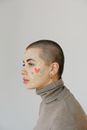 Side view of serious young woman in casual outfit and colorful heart stickers on face standing against gray background and looking away