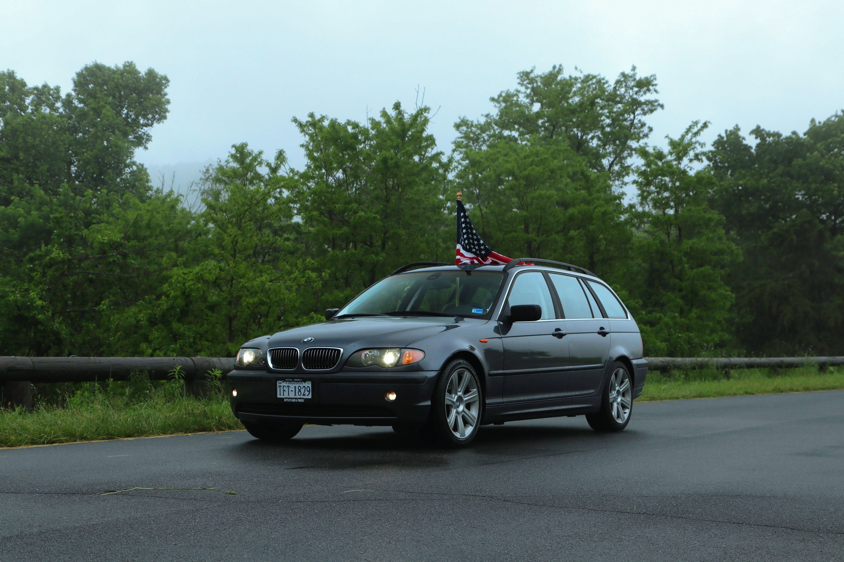 A BMW wagon carrying an American flag navigates a scenic, tree-lined road.