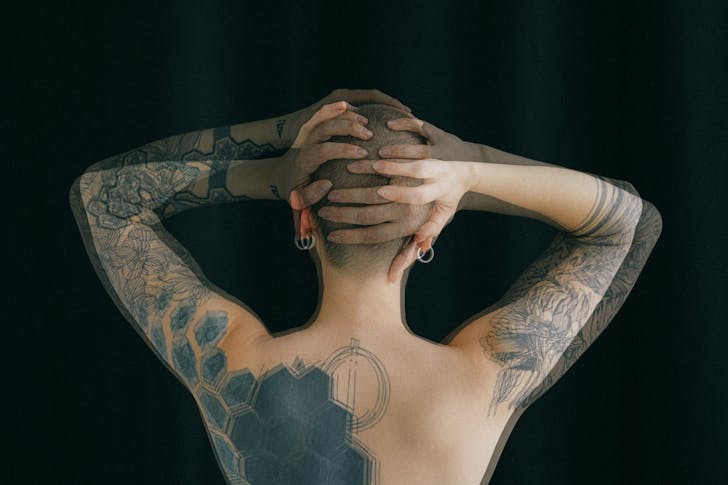 Back view of faceless topless female with tattoos on arm and back standing against black background and holding hands behind head in long expose