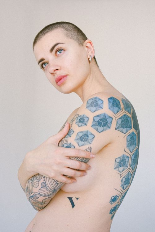 Free From below of slim naked young woman with many tattoos on body standing against gray background looking up and hugging shoulders Stock Photo