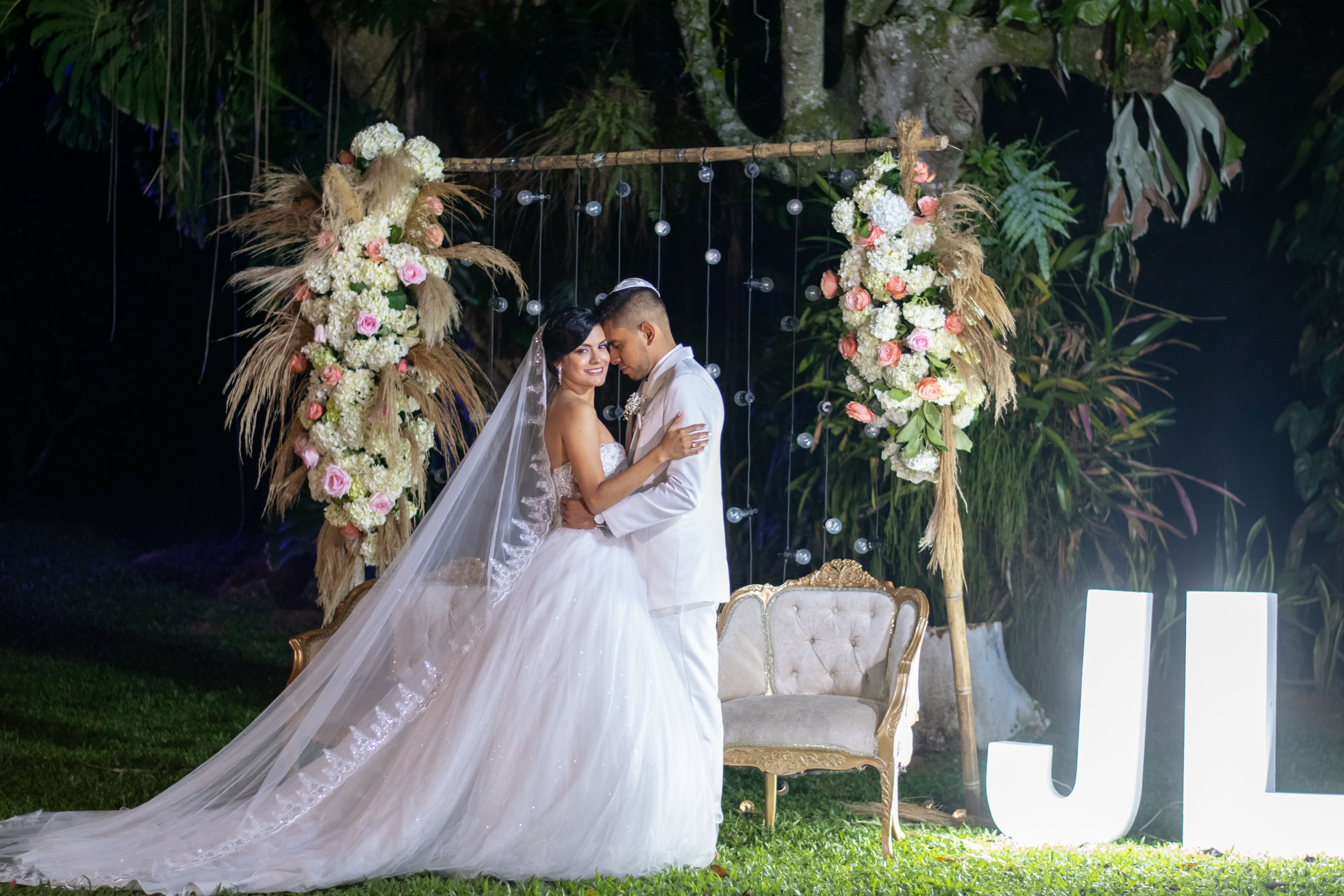 A newlywed couple embraces under floral decorations in a romantic nighttime outdoor setting.