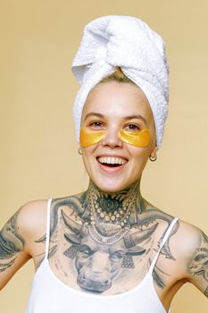 Young joyful female in casual wear with creative tattoos on body standing on beige background with bath towel on head and eye patch under eyes and looking at camera