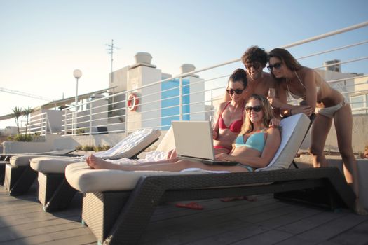 Group of happy young friends in swimwear browsing laptop and looking at screen while resting together on loungers near swimming pool