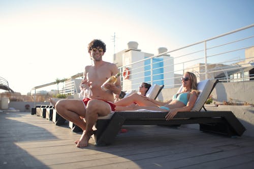Free Smiling guy in swimwear and sunglasses sitting on lounger near girlfriend holding bottle of white wine in one hand and four glasses in another and looking at camera while chilling with friends Stock Photo
