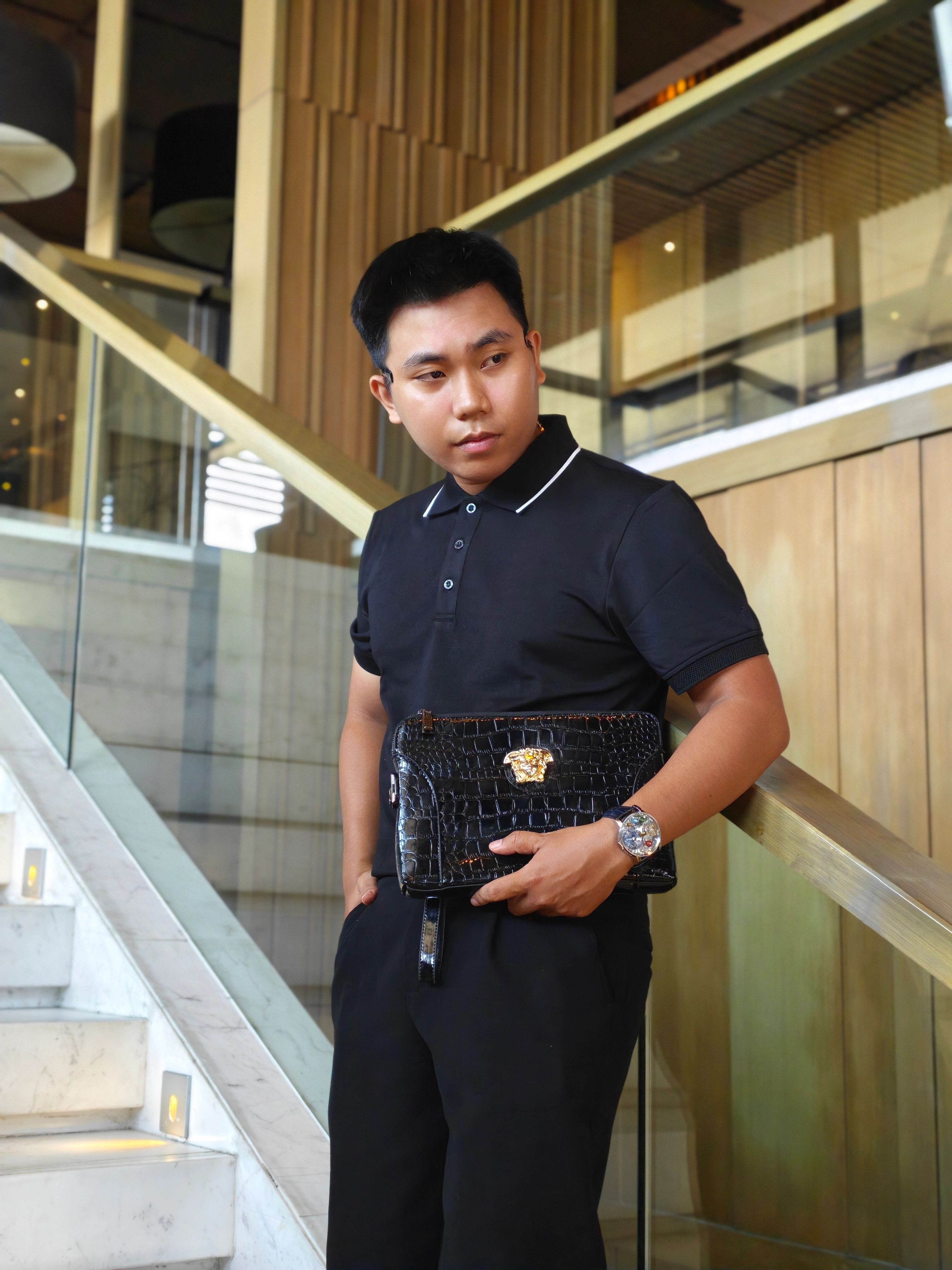 Homem com roupa preta segurando uma bolsa clutch de luxo indoors em uma escadaria.