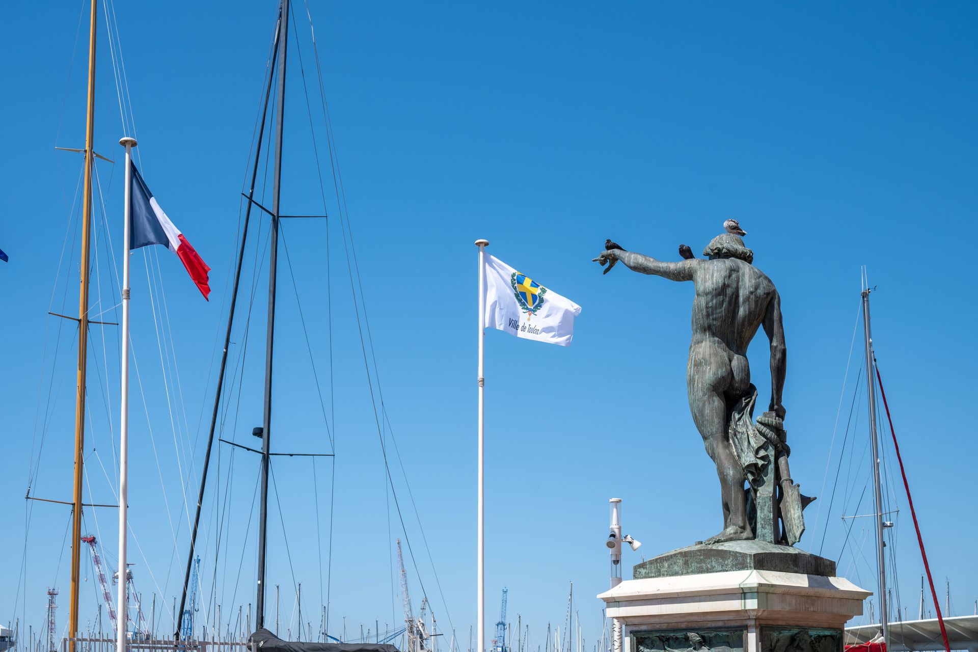 A statue overlooks sailboat masts with flags in Toulon Harbor, France under a clear blue sky.