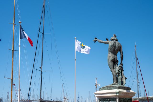 A statue overlooks sailboat masts with flags in Toulon Harbor, France under a clear blue sky.