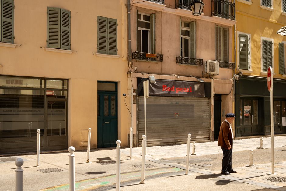 An elderly man walks past shuttered buildings on a sunny street in Toulon, France, capturing the calm urban scene.
