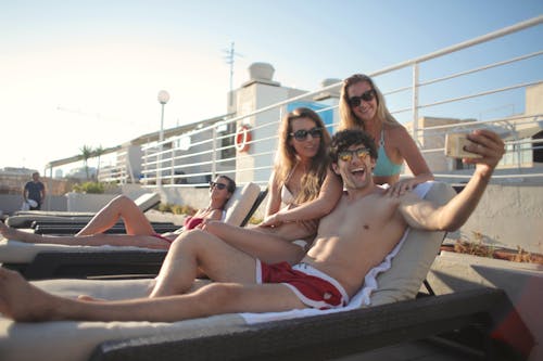 Free Group of young happy ladies and guy in swimsuits and sunglasses sitting on deck chair and taking selfie while chilling together during summer vacation Stock Photo