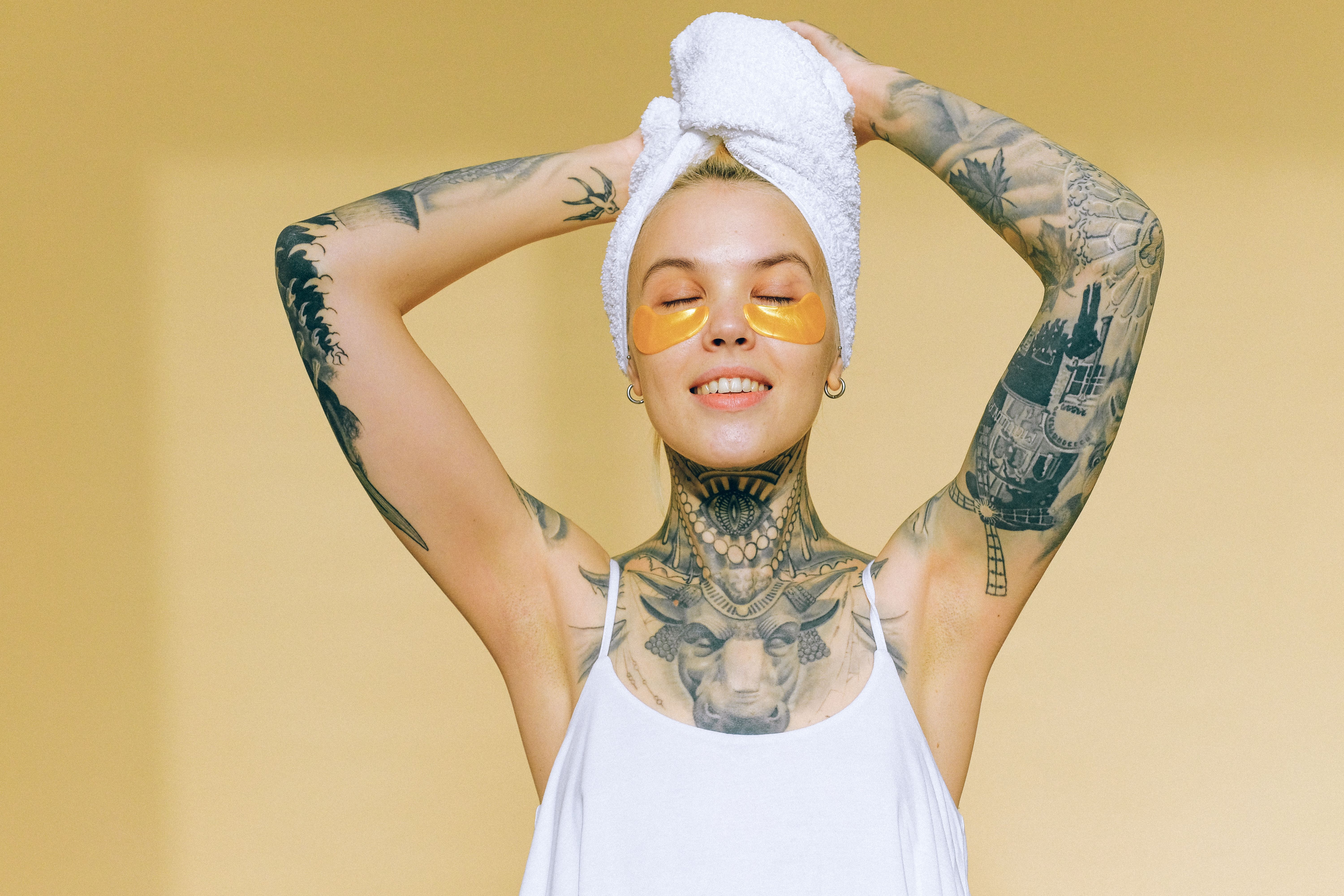 Tattooed woman enjoying spa day indoors with gold eye patches and towel on head.
