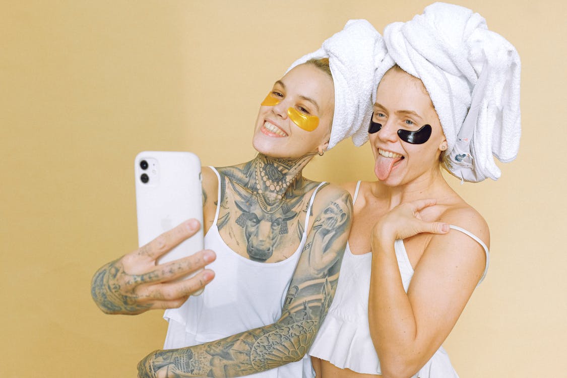 Viral tiktok beauty products being tested with optimistic girlfriends in white t shirts with straps bath towels on heads and eye patches standing on beige background and taking selfie on smartphone photo