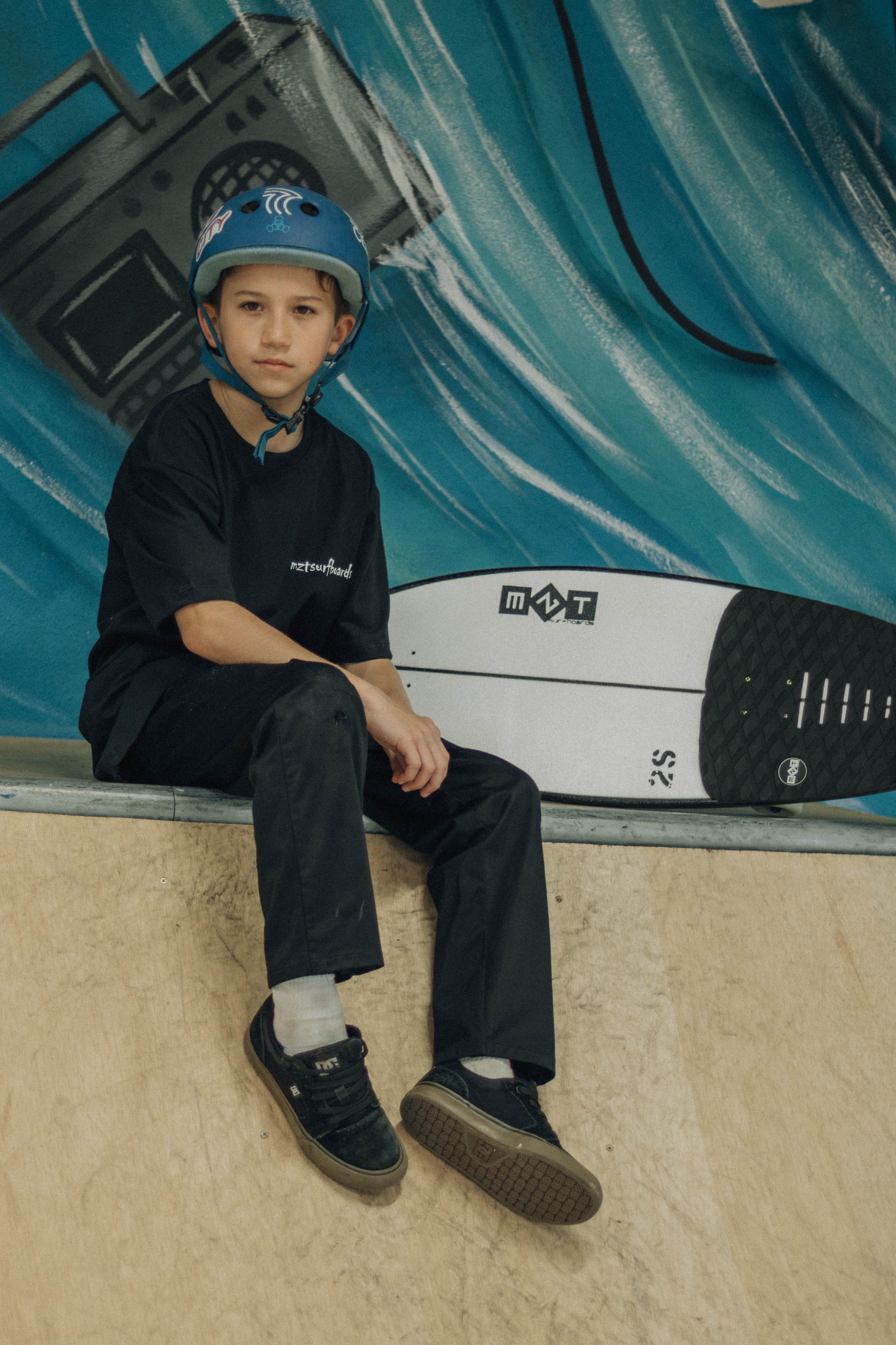 Young boy sitting in a skate park with a blue helmet and skateboard, ready for action.