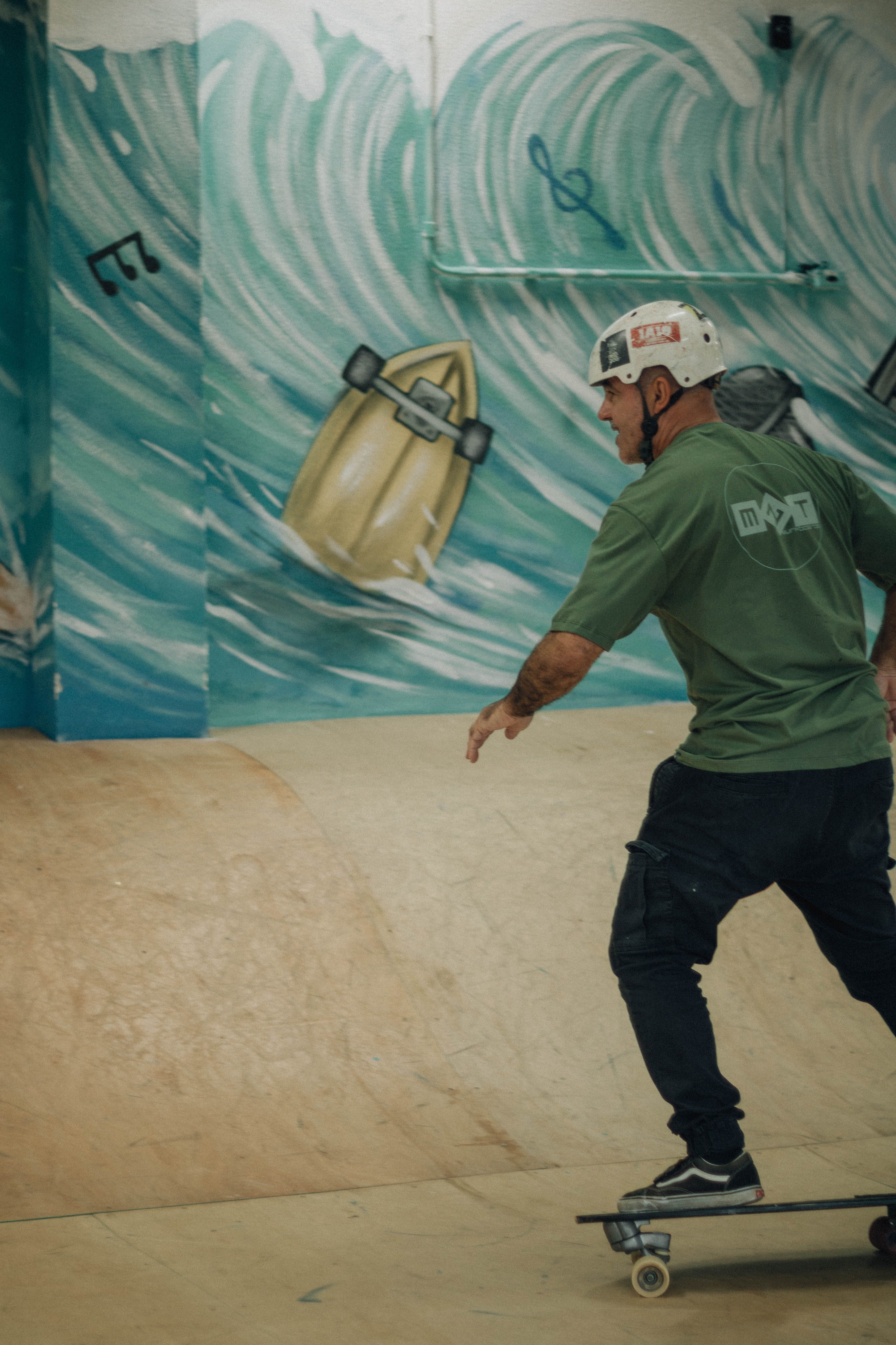 A skateboarder in a helmet rides indoors with a vibrant ocean wave mural backdrop.