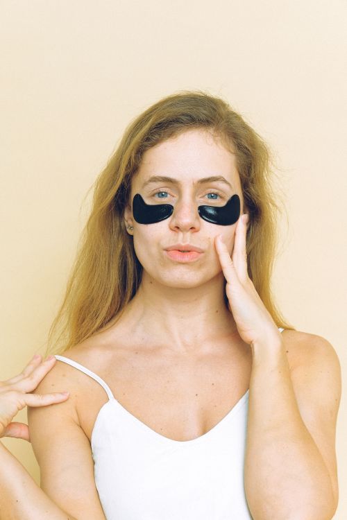 Free Positive young lady in white t shirt with straps in black eye patches under eyes standing against beige wall touching face with one hand and shoulder with another while looking at camera Stock Photo